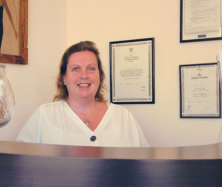 Profile Image of Sandra Whittle Standing in a clinical setting with framed qualification certificates on the wall nearby