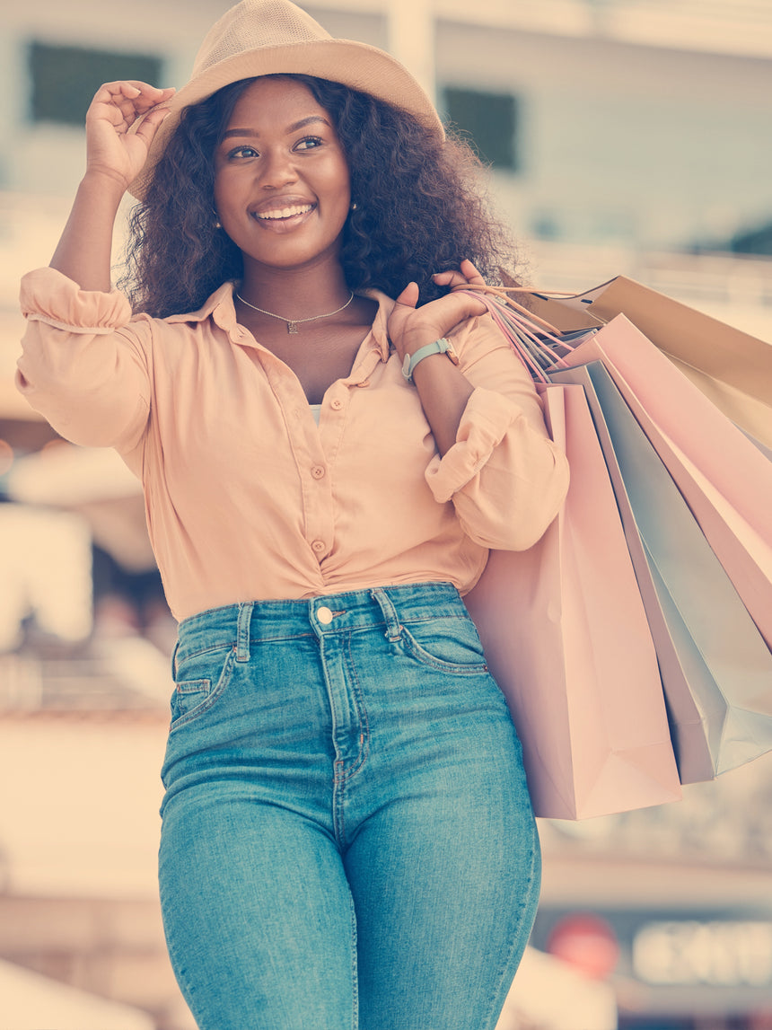 Woman holding shopping bags outdoors, wearing a beige hat and pink shirt with blue jeans.